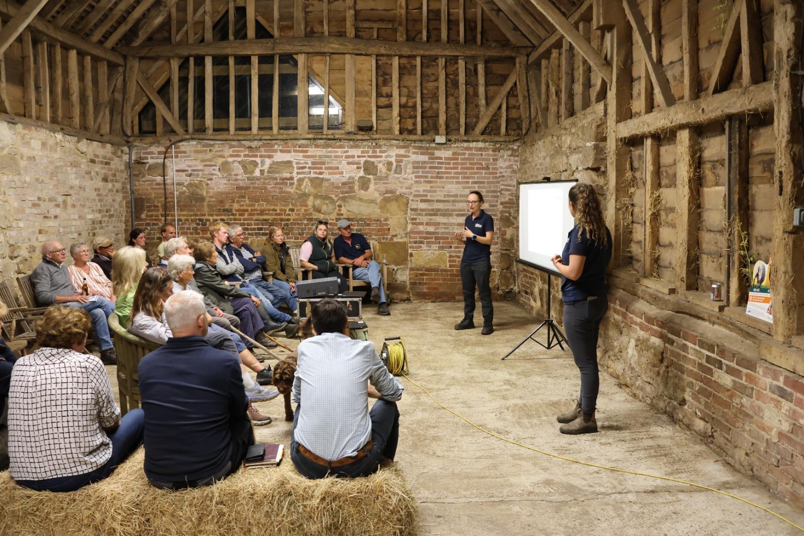 Farmers seated on straw bales and chairs in a timber-framed barn at Black Ven Farm listening to two Fairfield House vets giving a talk